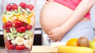 一位孕妇站在桌子旁边,桌子上放着新鲜水果和一个装满水果切块的搅拌机