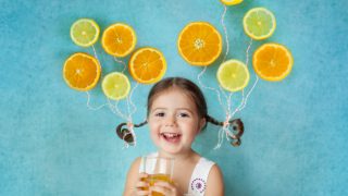 一个微笑的女孩拿着一杯果汁,她的马尾辫上绑着一半飞舞的柑橘,站在蓝色的背景下。