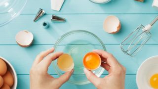 手拿蛋壳放在碗上,四周用打蛋器、鸡蛋、滚边袋和顶部,放在浅蓝色的木质表面上