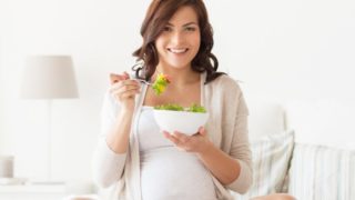 一位孕妇坐在一间白色的房间里,手里拿着一碗沙拉,看着镜头微笑着