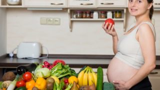 一位孕妇拿着一个苹果站在摆满蔬菜和水果的桌子前。