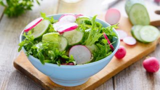 一个白色的碗,里面放着萝卜和黄瓜沙拉,放在木砧板上,后面是切好的蔬菜