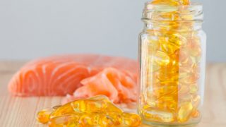 一个装满黄色药丸的玻璃瓶的特写,旁边放着更多的药丸,放在一个木头表面上,后面放着一块模糊的鱼肉。