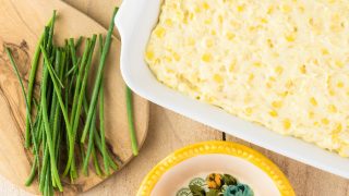 木桌上放着一盘未烤的奶油芝士玉米砂锅，旁边放着几碗奶酪和芦笋